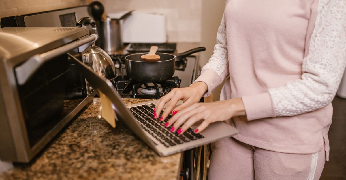 A woman in a pink sweater using a laptop in a modern kitchen, multitasking work and cooking.